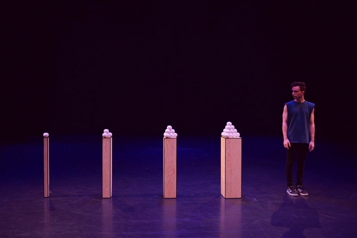 Performer Domenyk La Terra standing next to four tall plinths, each with a different number of juggling balls arranged in a pyramid on top of it.