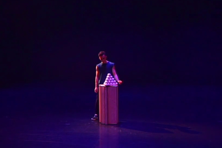 Performer Domenyk La Terra standing behind a single tall plinth, with 35 juggling balls arranged in a single pyramid on top of it.
